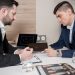 Two businessmen in suits discussing cryptocurrency trends using digital devices in an office setting.